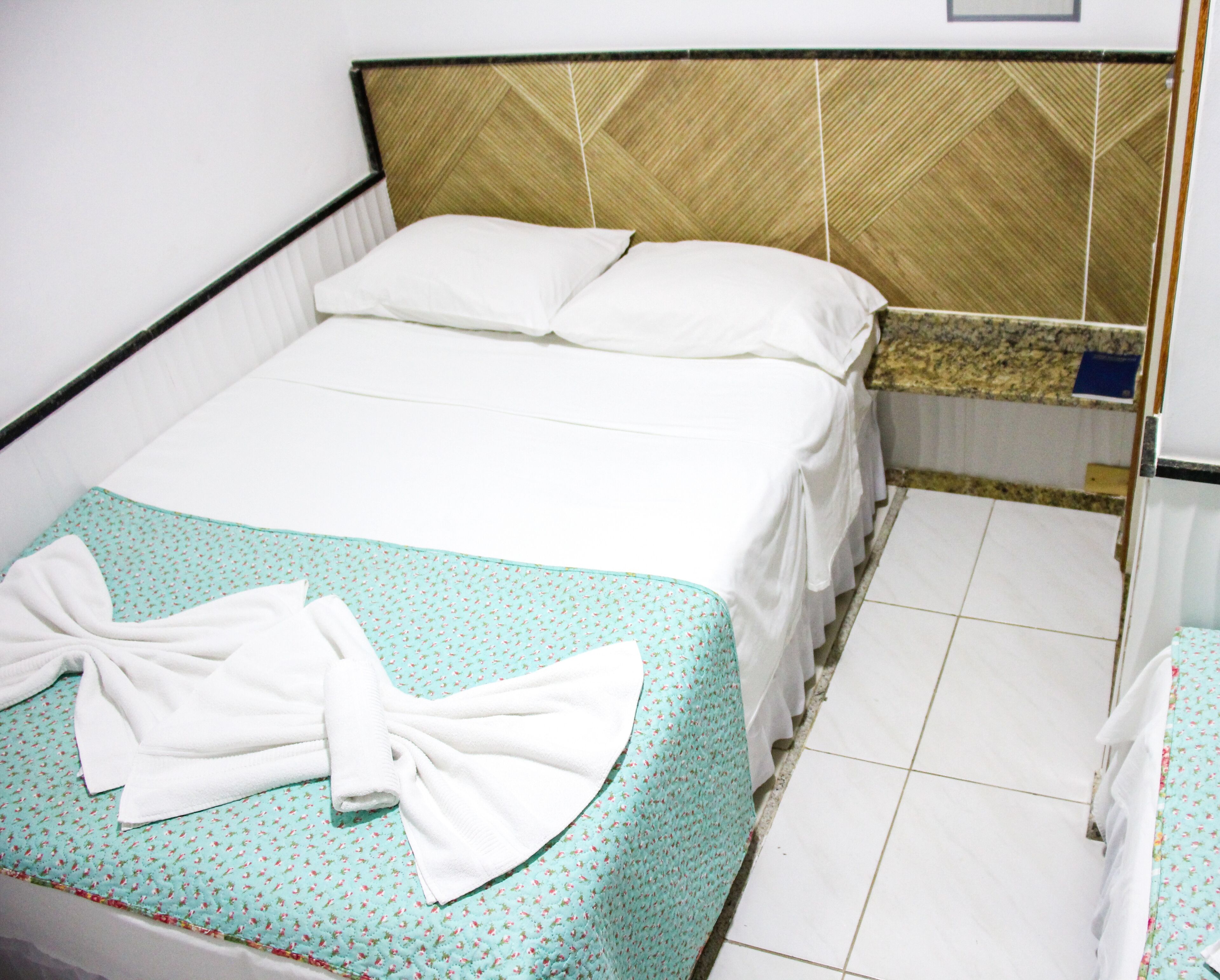A neatly made bed with a green and white patterned bedspread, two pillows, and a white towel folded on the bed.