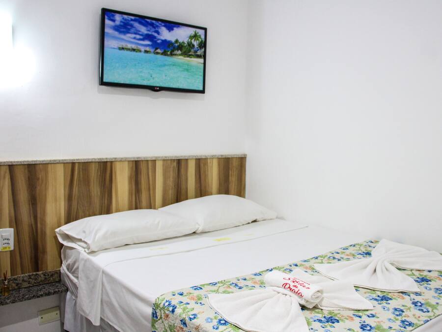 A hotel room with a bed, a wooden headboard, a wall-mounted TV displaying a beach scene, and a floral bedspread.