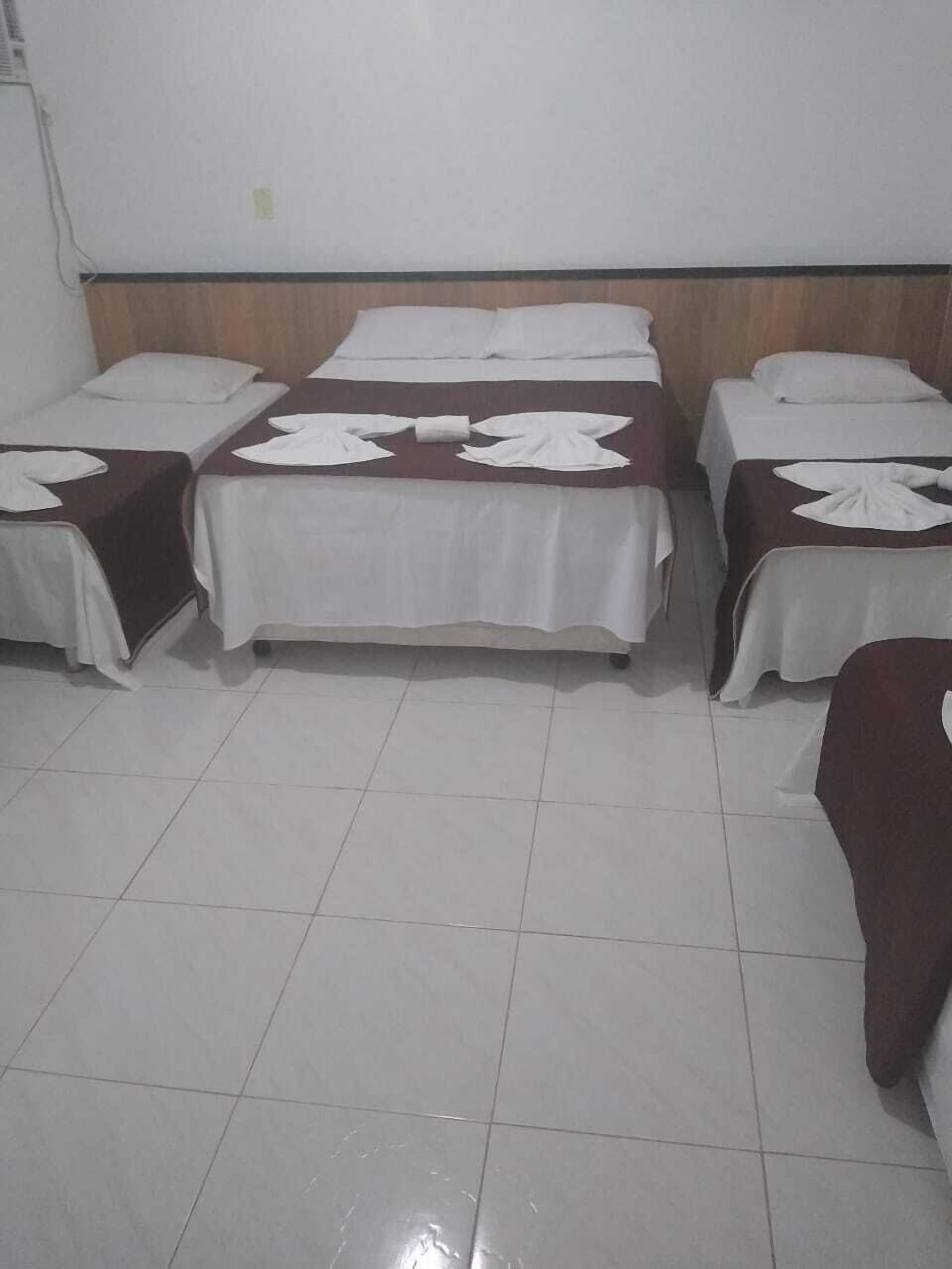 A hotel room with three single beds, each with a white bedspread and a towel folded at the foot. The room has a tiled floor and a wooden headboard.