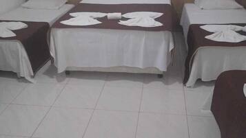 A hotel room with three single beds, each with a white bedspread and a towel folded at the foot. The room has a tiled floor and a wooden headboard.