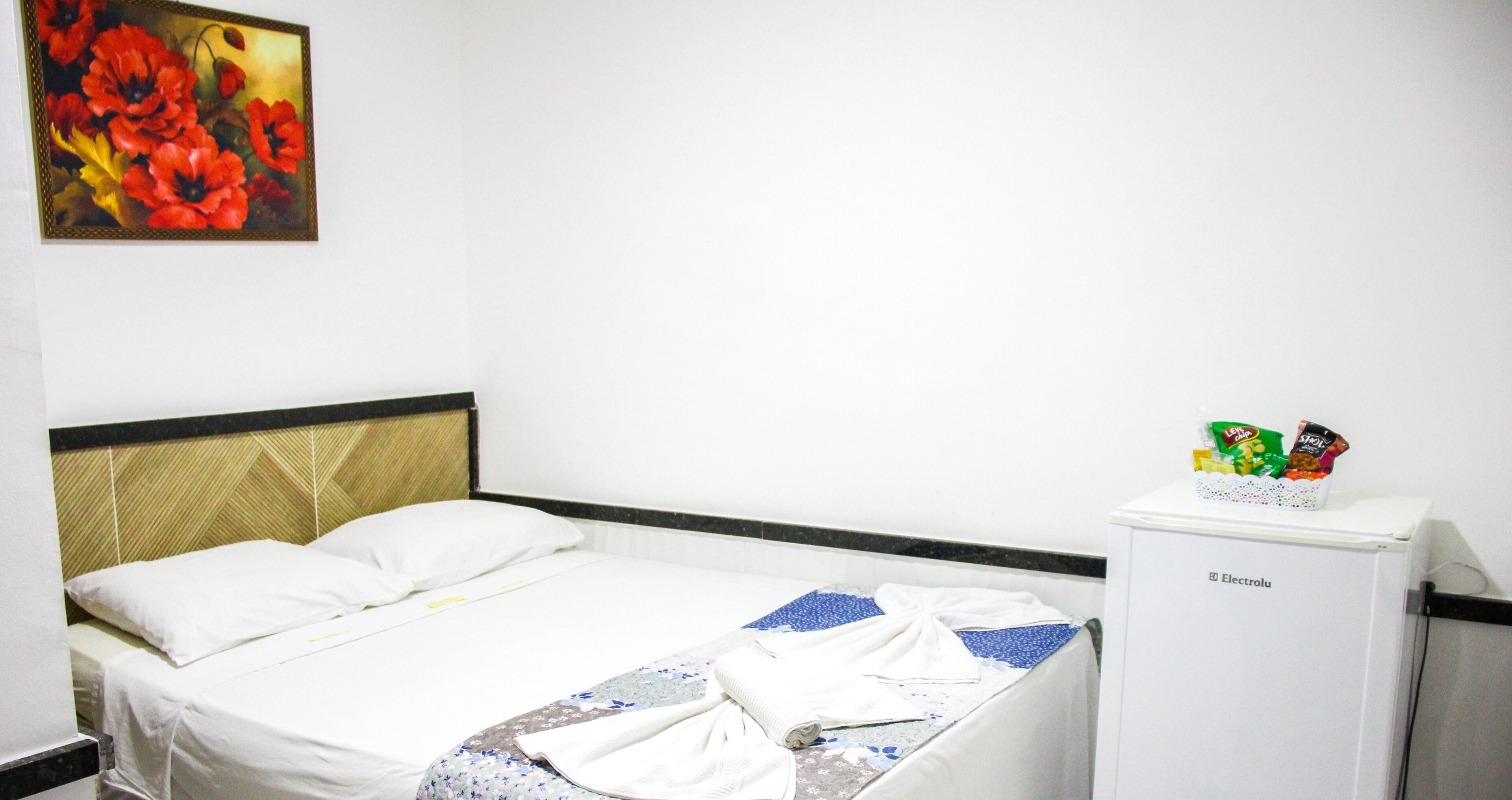 Una habitación de hotel con una cama, un refrigerador pequeño y un cuadro de flores en la pared.
