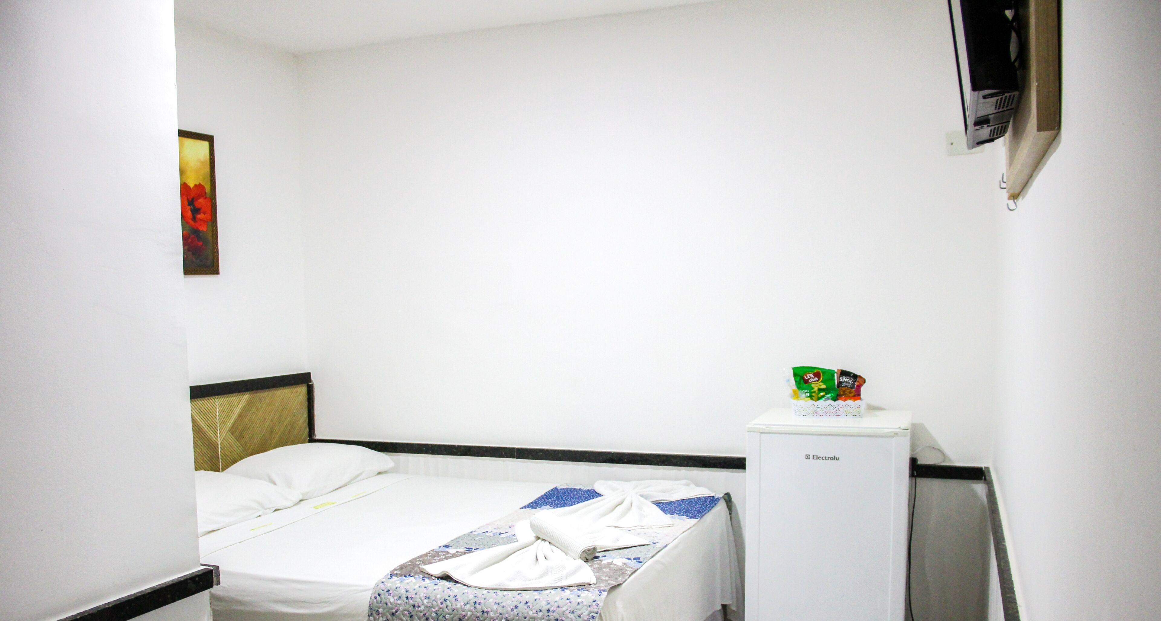 A hotel room with a bed, a small white refrigerator, and a wall-mounted TV.