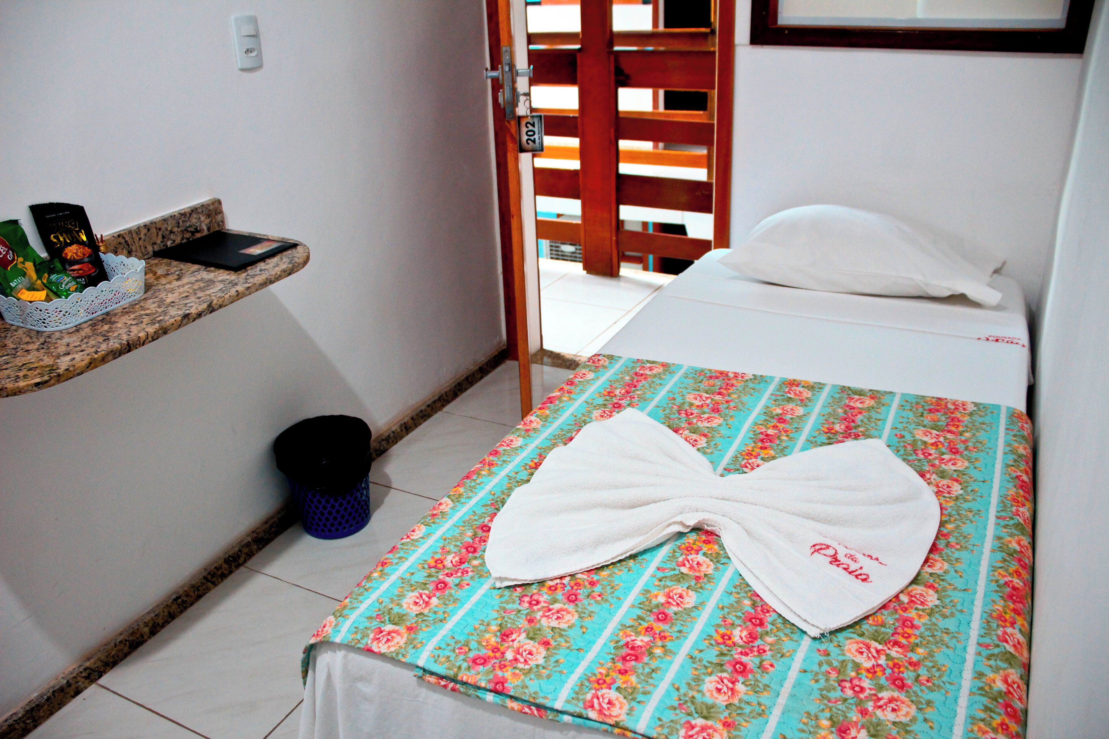 A hotel room with a bed, a small shelf with toiletries, and a door leading to another room.