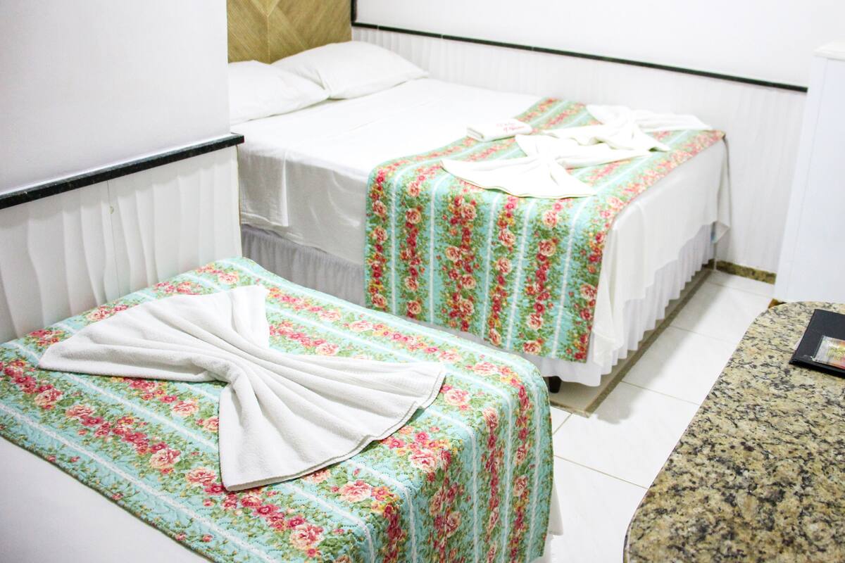 A room with two beds, one with a plain white bedspread and the other with a floral bedspread. There is a small table with a book on it.