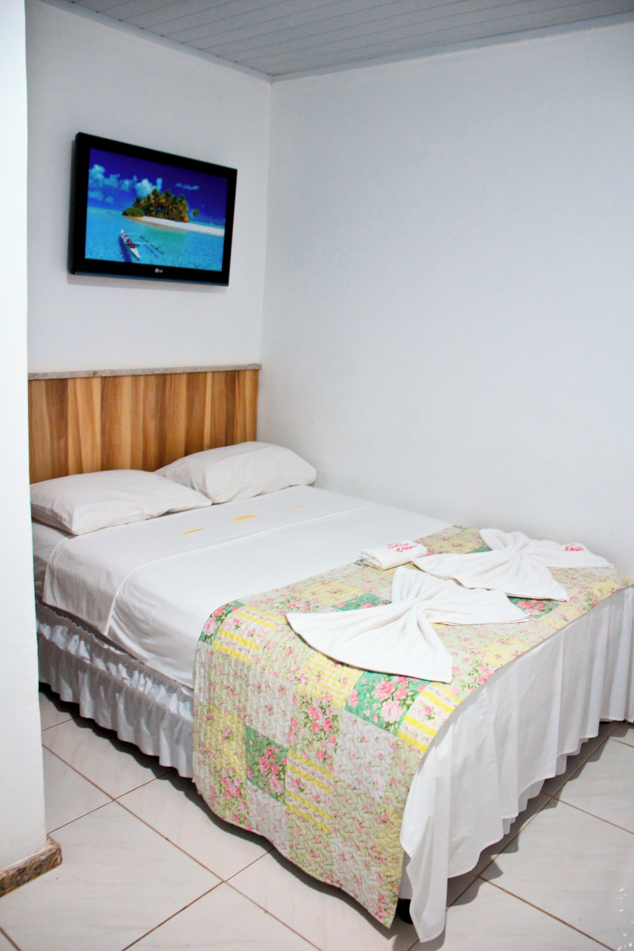 A neatly made bed with a floral bedspread and towels, a wooden headboard, and a television mounted on the wall displaying a tropical scene.