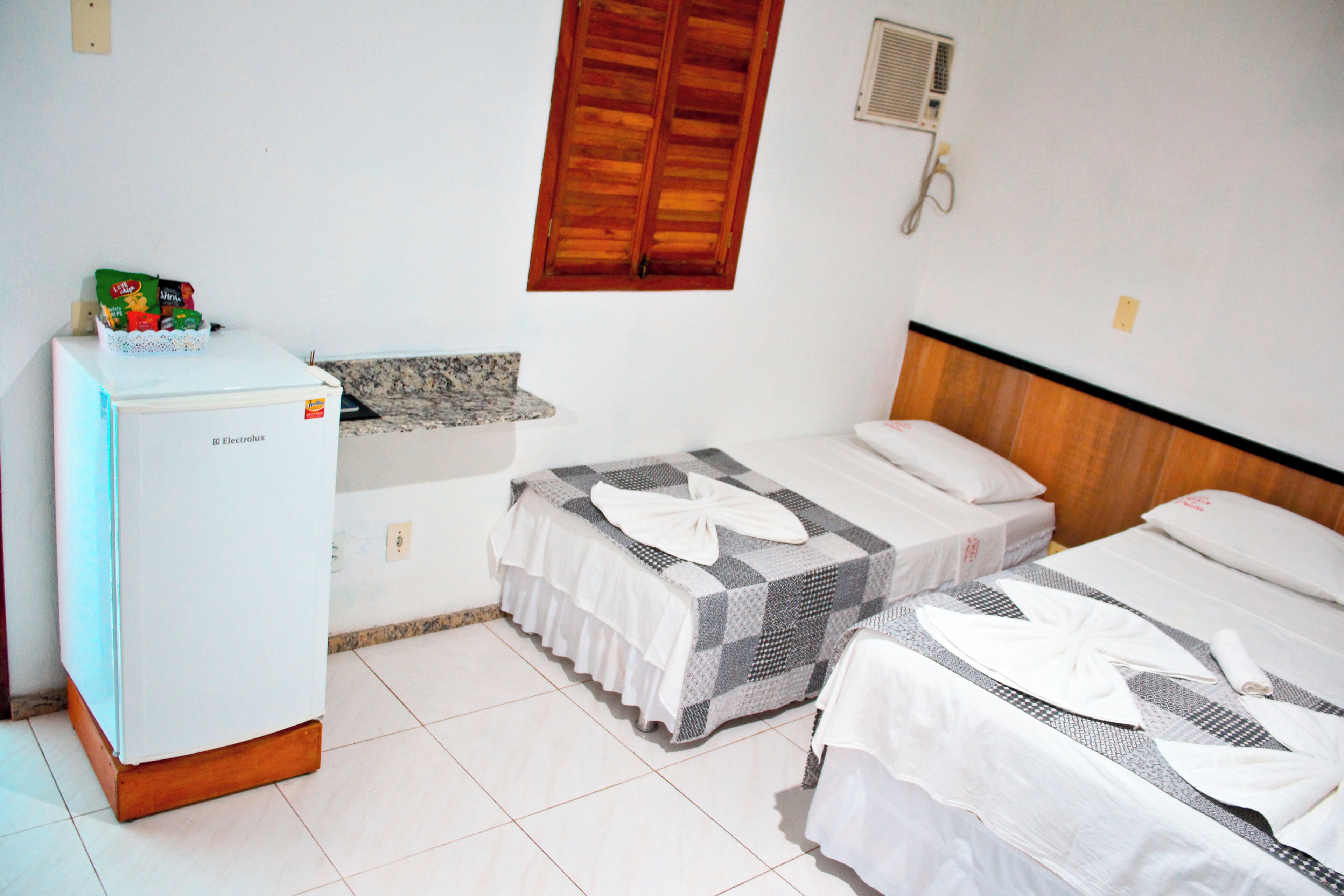 Habitación de hotel con dos camas, un refrigerador, una ventana con contraventanas de madera y aire acondicionado instalado en la pared.