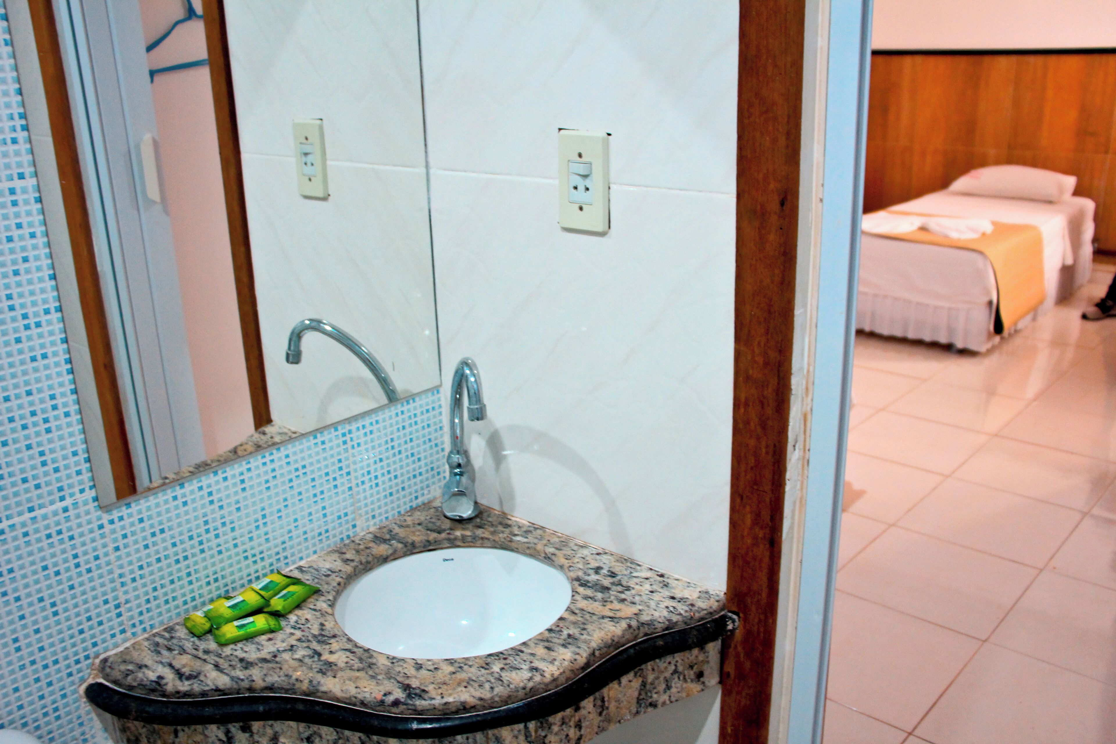 A bathroom with a granite countertop sink, a mirror, and a tiled wall. There are two beds visible in the background.