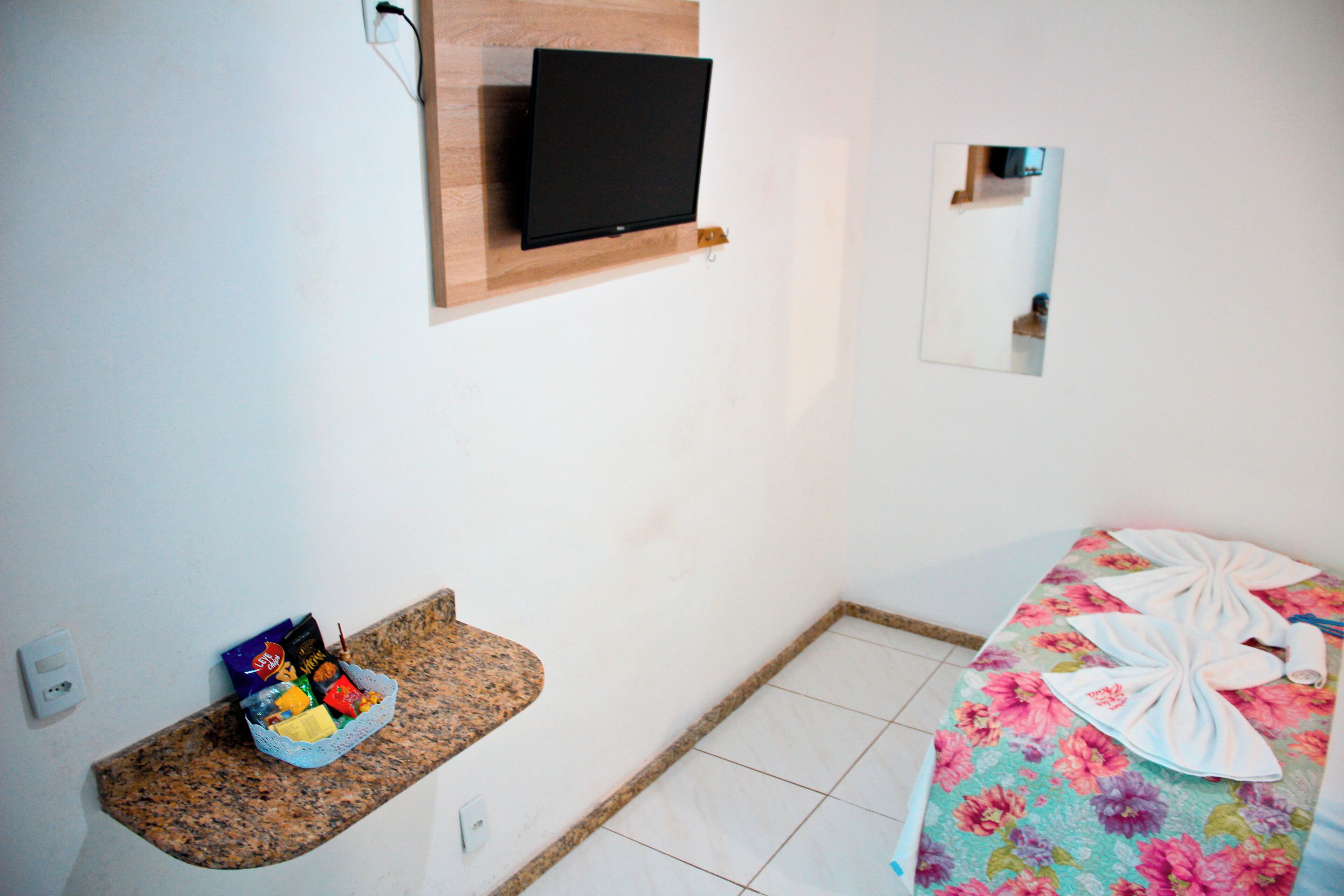 Habitación de hotel con televisor de pantalla plana en la pared, una pequeña estantería con aperitivos, una cama con colcha estampada y un espejo en la pared.