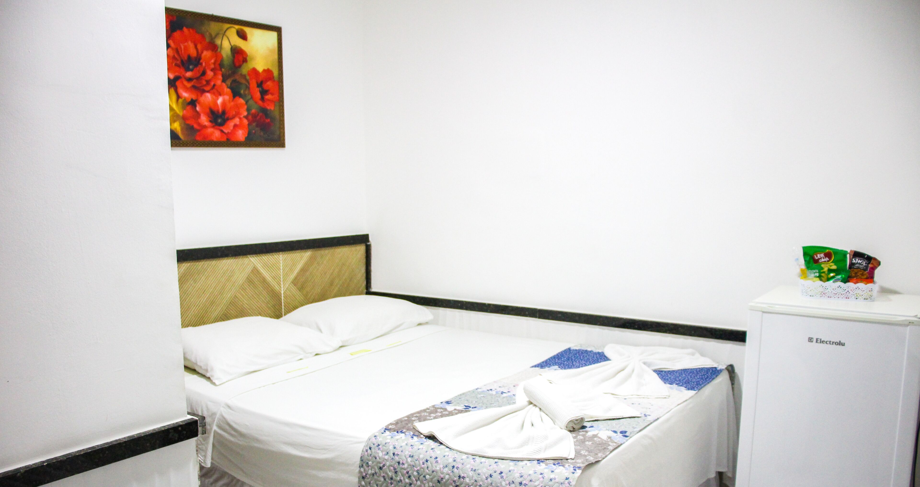 A hotel room with a bed, a headboard, a wall-mounted painting, and a small refrigerator.