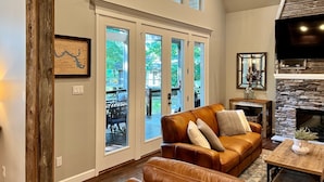 A spacious living room with a leather sofa, a stone fireplace, and large windows offering a view of the outdoors. - Castaway Cove-Hot Tub, Boat Dock + Theater Room (Hot Springs)