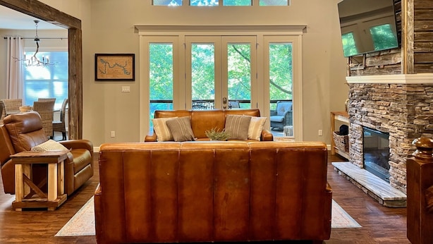 A spacious living room with a leather sofa, a stone fireplace, and large windows overlooking a deck. - Castaway Cove-Hot Tub, Boat Dock + Theater Room (Hot Springs)