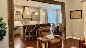 A spacious living area with a leather armchair, a wooden coffee table, and a dining area with wicker chairs. A kitchen with a brick backsplash and wooden cabinets is visible through an open doorway. - Castaway Cove-Hot Tub, Boat Dock + Theater Room (Hot Springs)