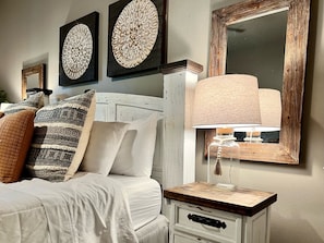 A bedroom with a white bed, patterned pillows, a wooden nightstand with a lamp, and two wall-mounted decorative mirrors.