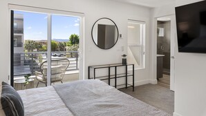 A modern bedroom with a large bed, a round mirror, a small table, and a balcony with a view of the city. - Perfect Escape - Brand New St. George Townhome (St. George)