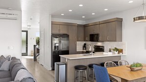 A modern kitchen with a central island, stainless steel appliances, and a dining area with a wooden table and bar stools. - Perfect Escape - Brand New St. George Townhome (St. George)