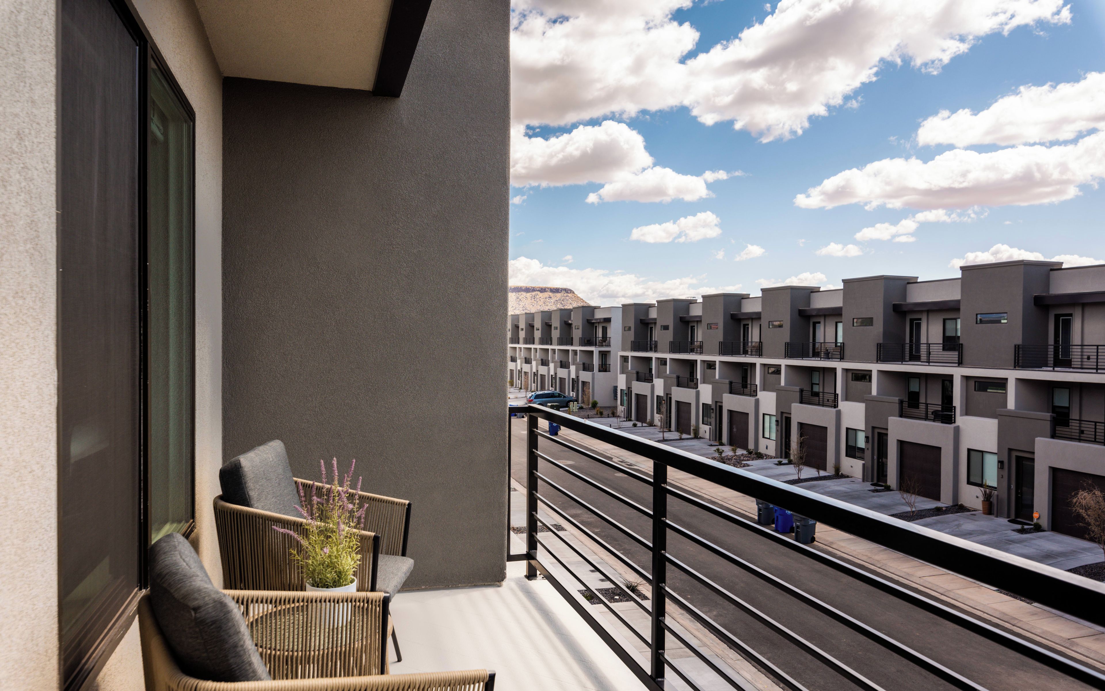 Ein Balkon mit Korbstühlen und einem kleinen Topfpflanzen, mit Blick auf eine Reihe moderner Gebäude und einen klaren Himmel.
