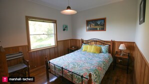 A bedroom with a bed, a bench, a nightstand, a lamp, a window, and a painting on the wall. - Moorhead Beach Cottage - Wallaga Lake Rd 60 (Bermagui)