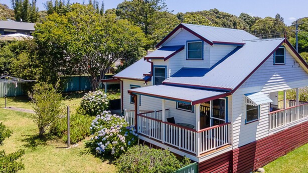 A two-story house with a porch and a small deck, surrounded by greenery and trees. - Moorhead Beach Cottage - Wallaga Lake Rd 60 (Bermagui)