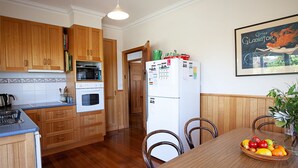 A kitchen with wooden cabinets, a microwave, oven, and a refrigerator with magnets. There is a dining table with chairs and a fruit bowl. - Moorhead Beach Cottage - Wallaga Lake Rd 60 (Bermagui)