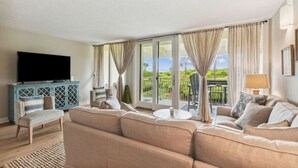 A living room with a large flat-screen TV, a blue cabinet, a beige sofa, and a balcony with a view of greenery.