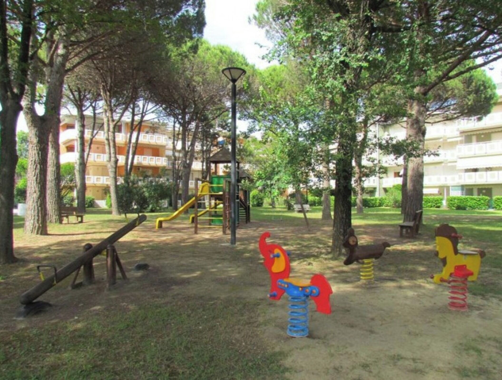 A playground with swings, slides, and climbing structures surrounded by trees and a building in the background.
