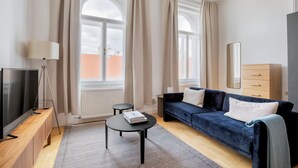 A modern living room with a dark blue sofa, a small round table, a television on a wooden stand, and a large window with curtains.
