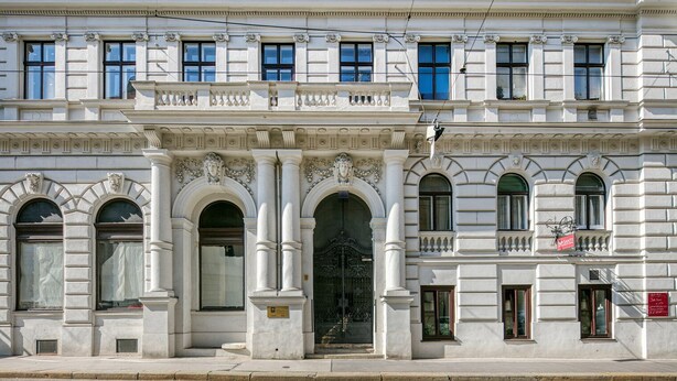A classical building with arched windows, a central entrance with a decorative door, and a balcony with a sign.