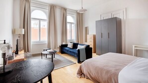 A modern hotel room with a bed, a sofa, a desk with a lamp, and a wardrobe.