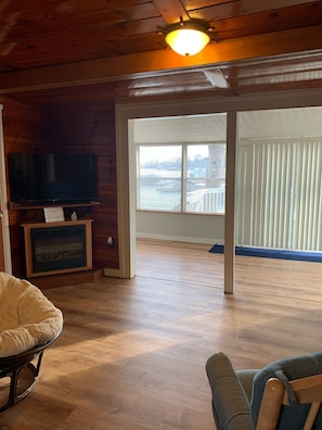 A room with wooden walls, a television, a fireplace, and a large window with blinds.