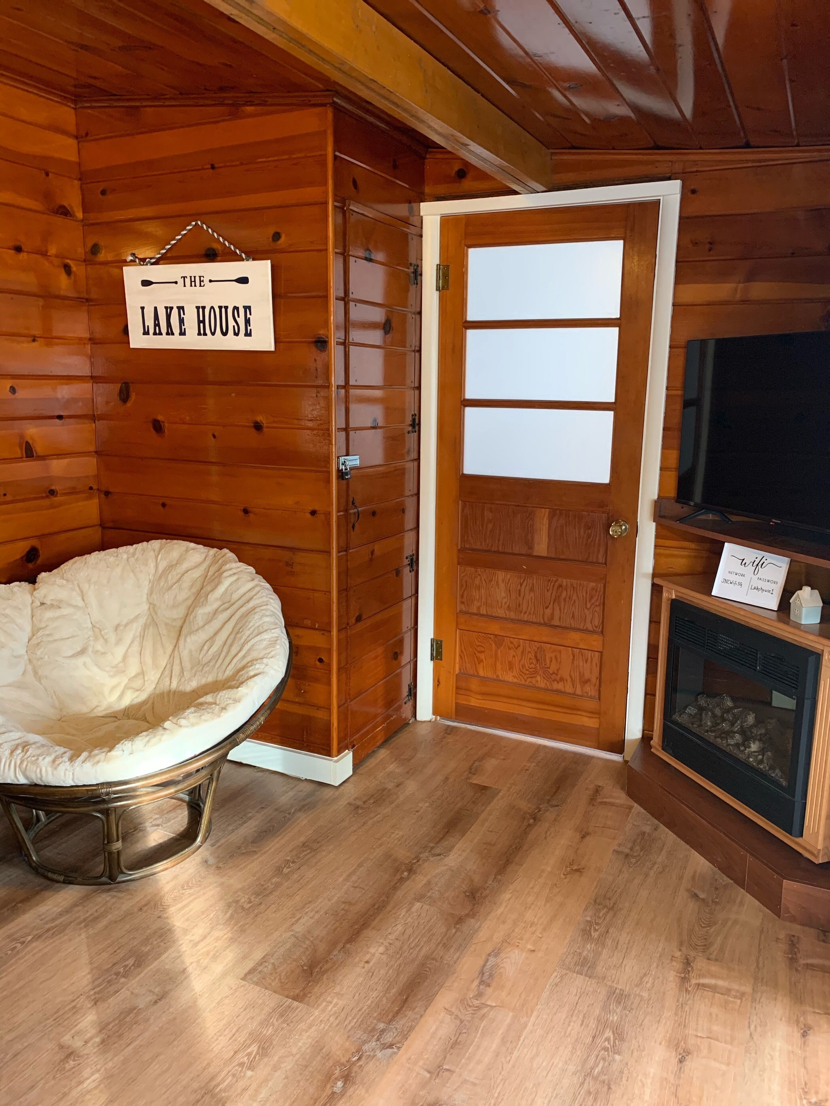 Un intérieur en bois avec une enseigne indiquant « The Lake House », un fauteuil blanc rembourré, une porte en bois et une cheminée.