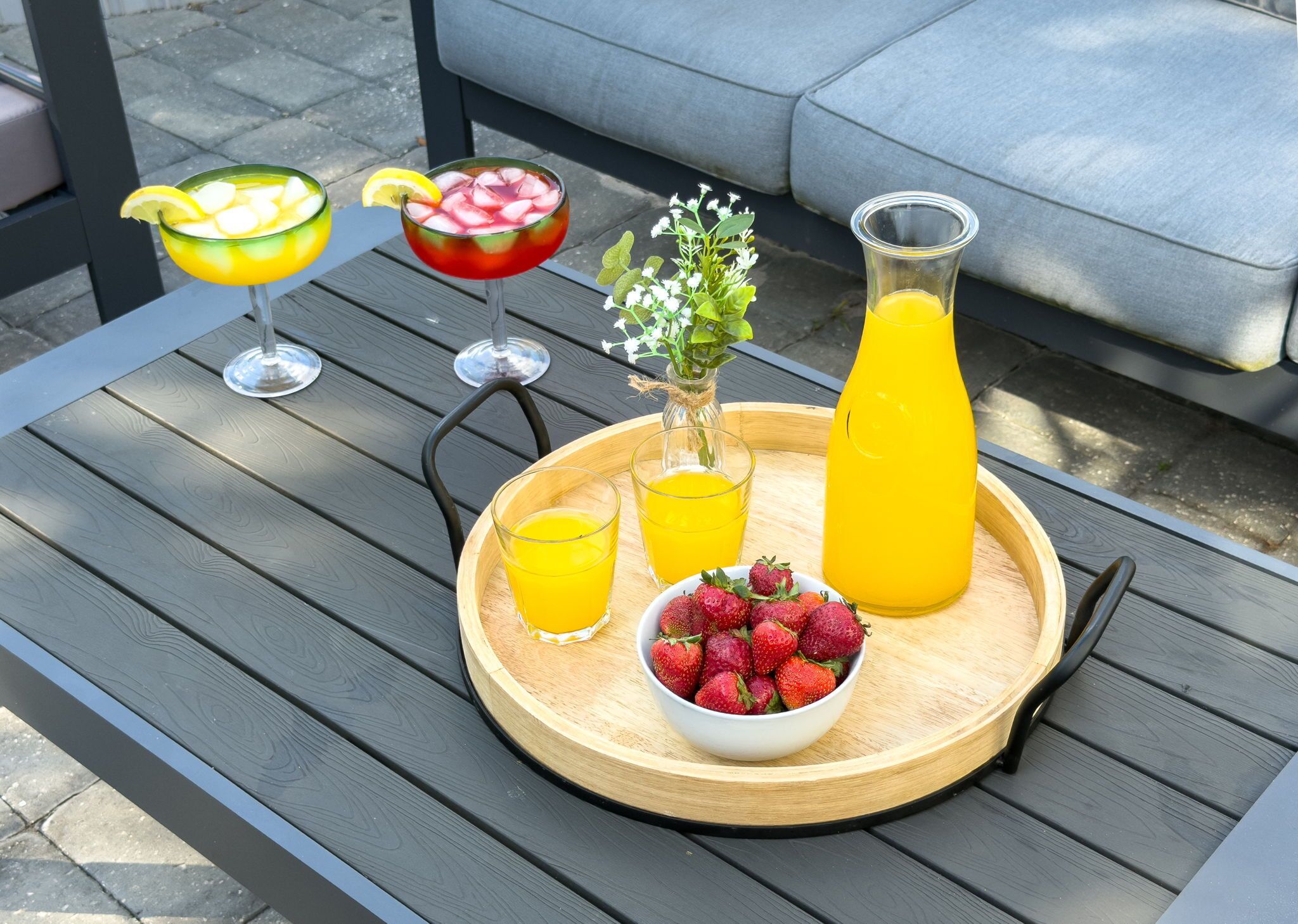 A wooden tray with drinks, a pitcher, and a bowl of strawberries on an outdoor table.