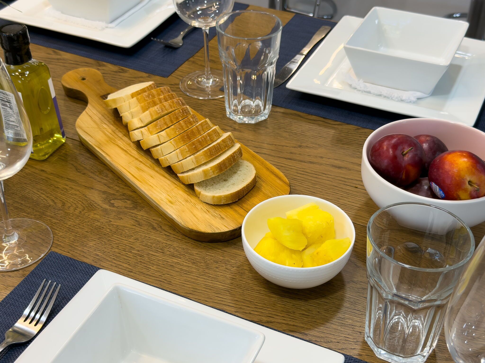 Una mesa de comedor con una tabla de madera que sostiene pan en rodajas, un tazón de trozos de piña y un tazón de ciruelas.