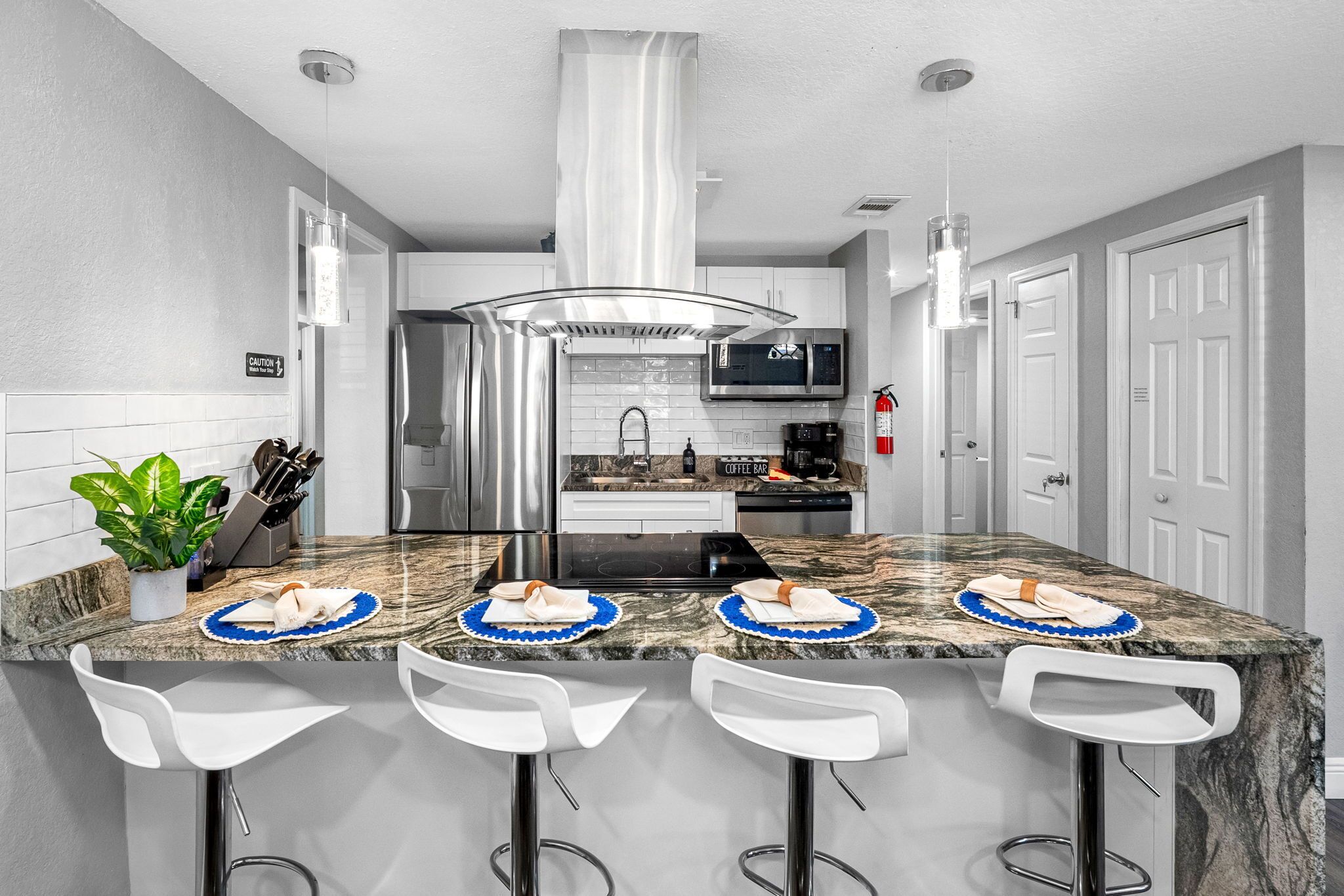 A modern kitchen with a central island, bar stools, stainless steel appliances, and a potted plant.