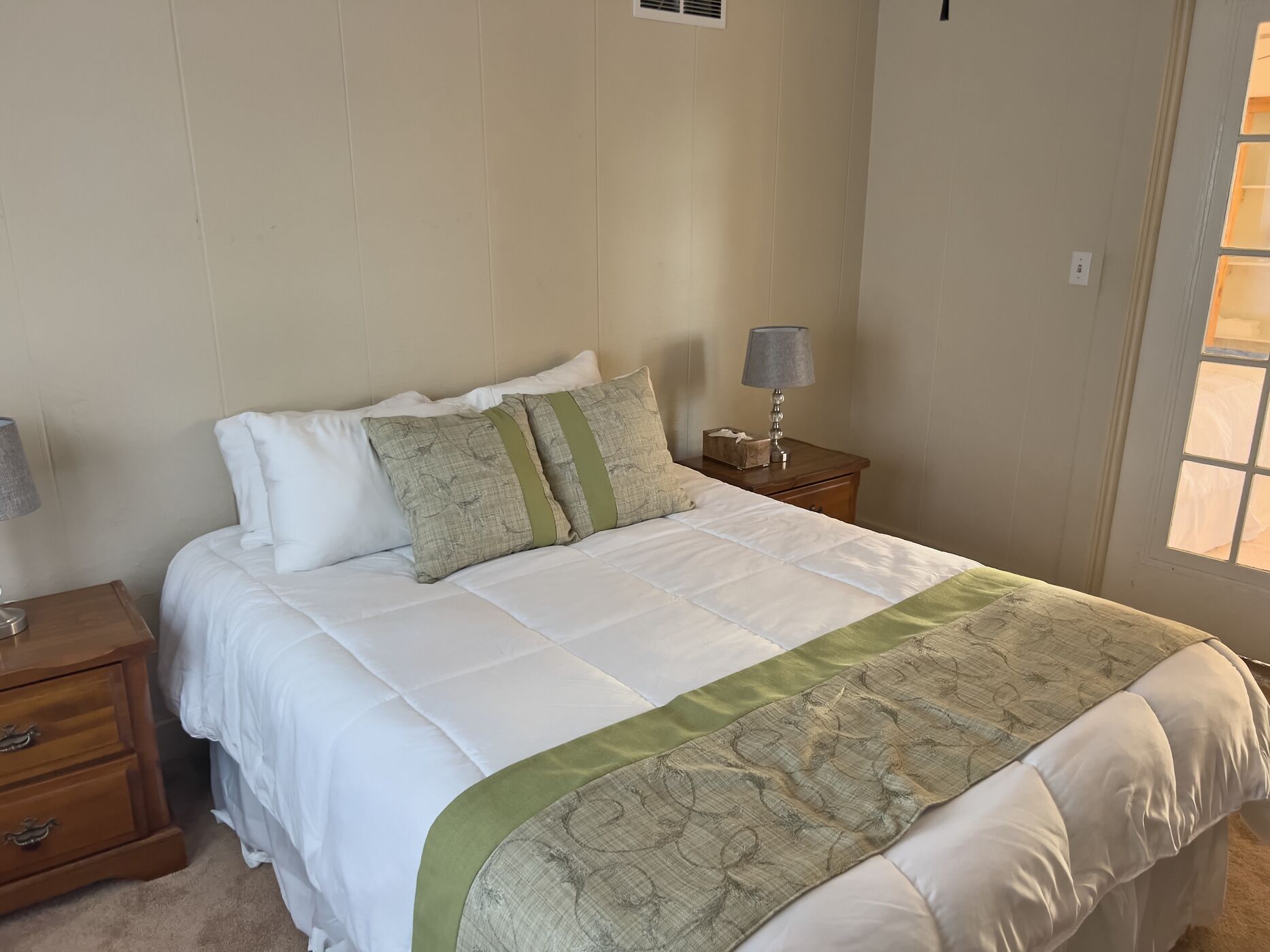 A neatly made bed with white bedding and a green accent throw, a bedside lamp, and a wooden nightstand with drawers.