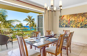 A dining area with a glass table, wooden chairs, and a view of the ocean and palm trees.