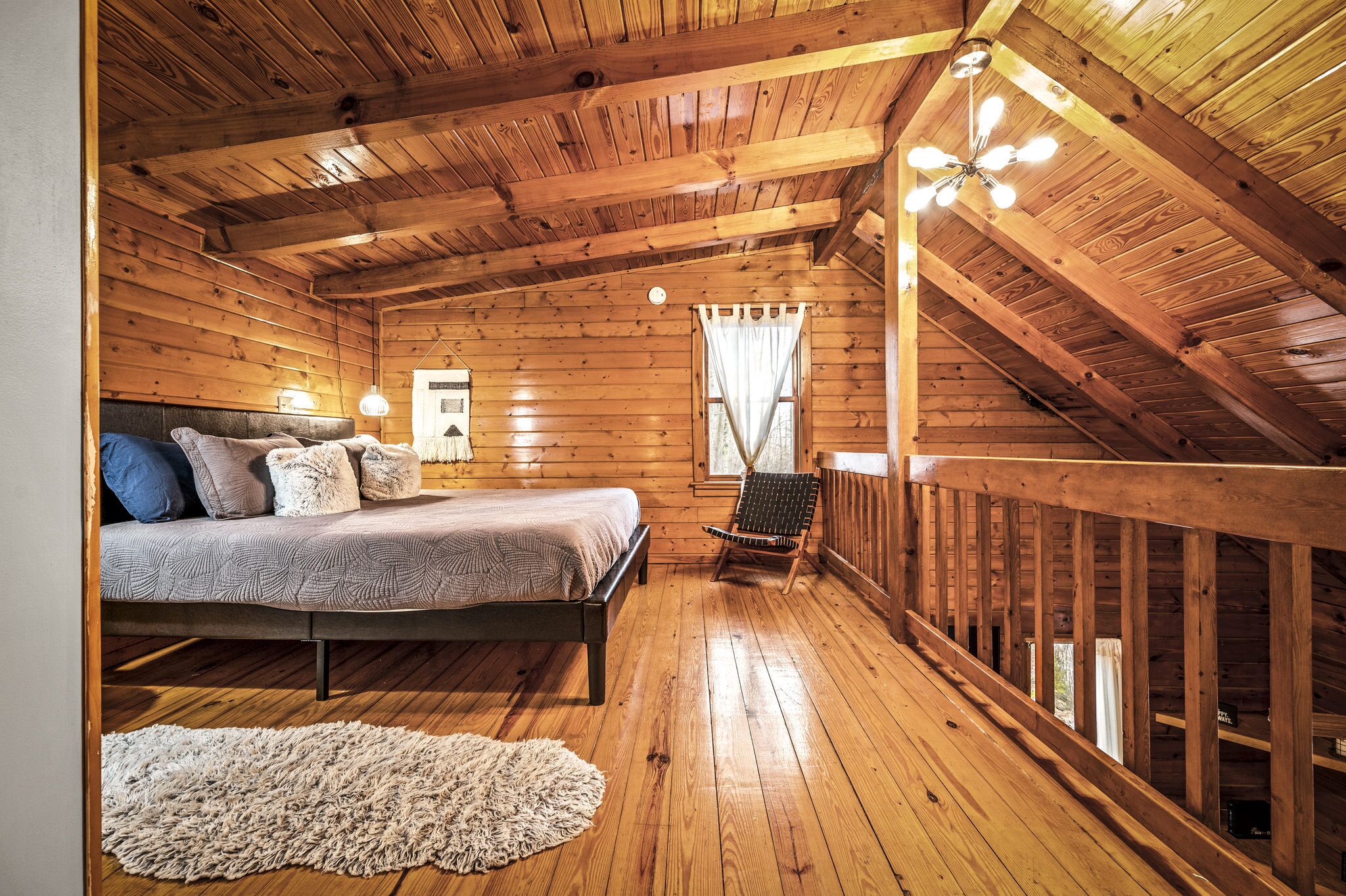 A wooden cabin bedroom with a bed, a chair, a small table, and a window with curtains.