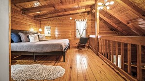 A wooden cabin bedroom with a bed, a chair, a small table, and a window with curtains.