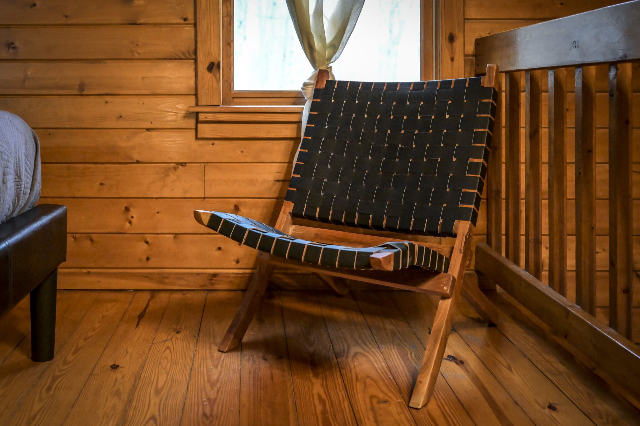 A wooden cabin interior with a single wooden chair with a black and green woven backrest, a wooden bench, and a window with a curtain.