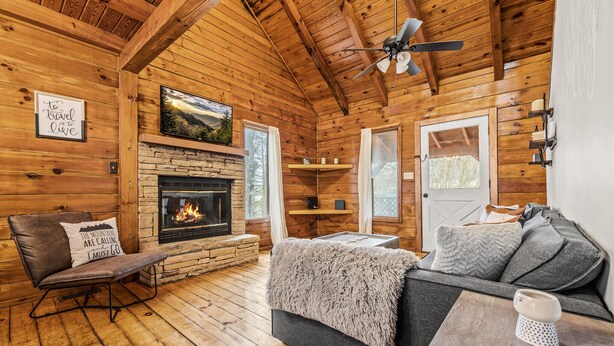 A cozy cabin interior with a stone fireplace, wooden walls, a leather chair, a gray sofa, and a ceiling fan.