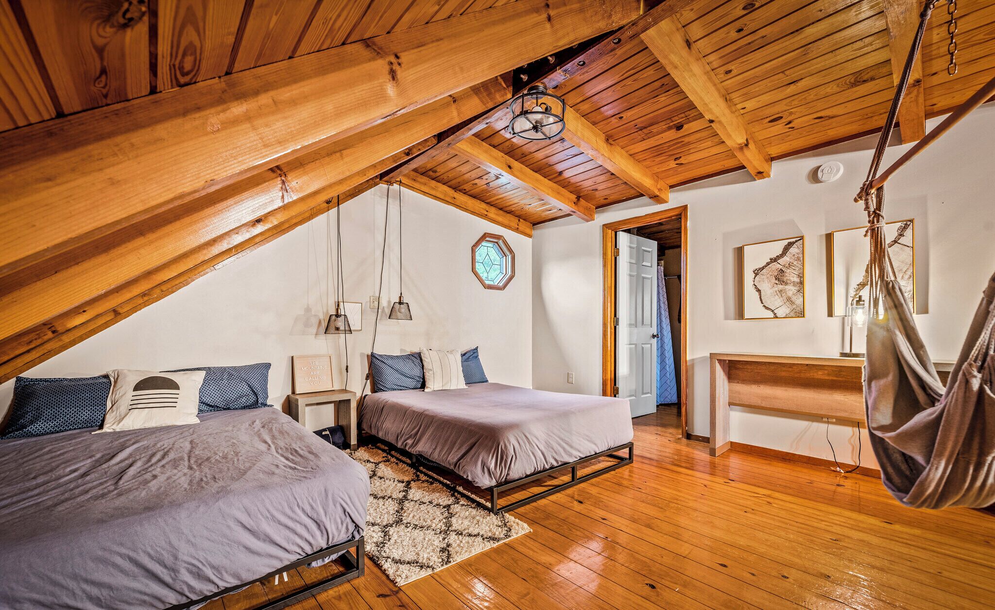 A loft bedroom with two beds, wooden beams, a hanging light fixture, and a view of the outside.