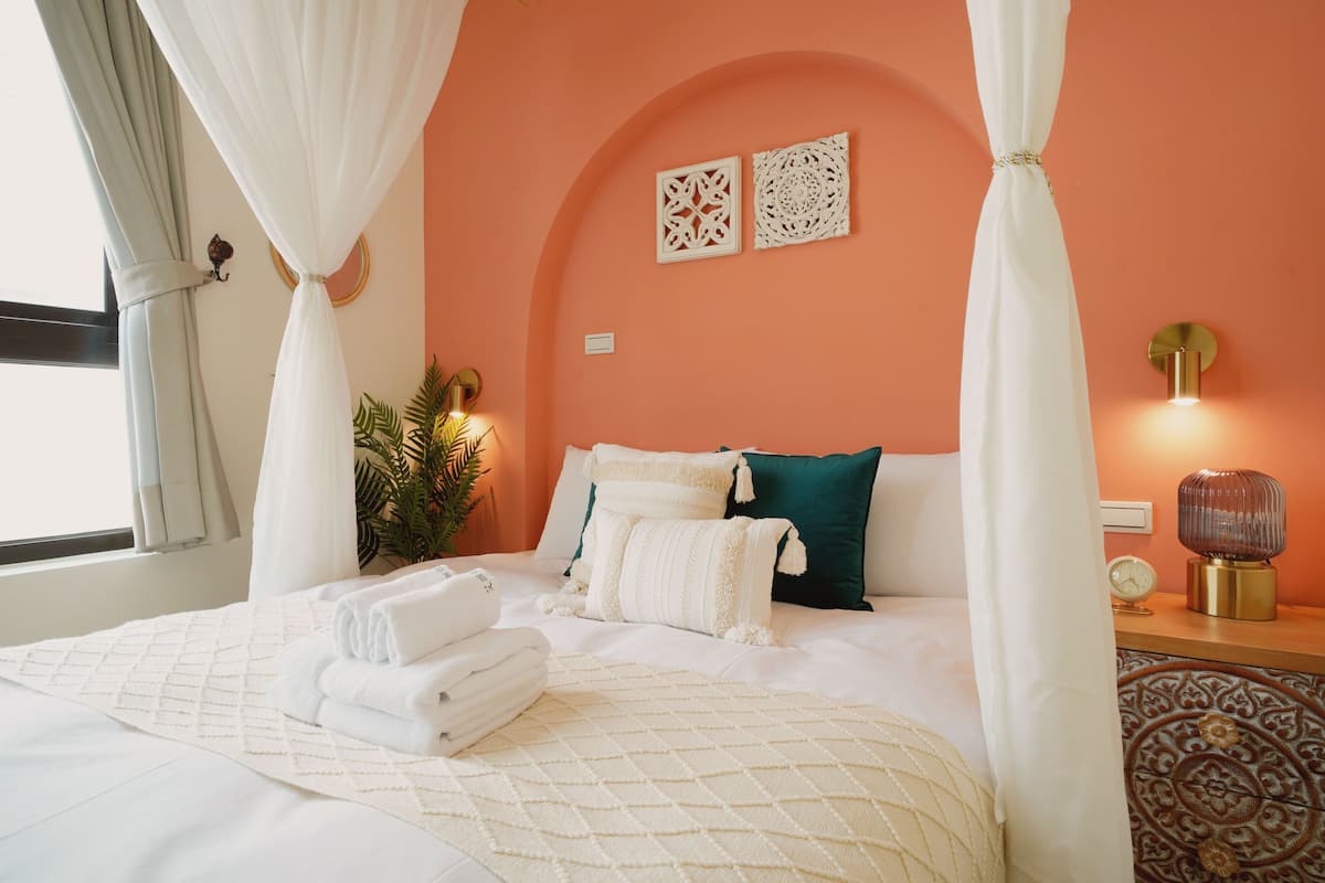 A neatly made bed with white linens and a quilted blanket, a bedside table with a lamp, and a wall-mounted clock.