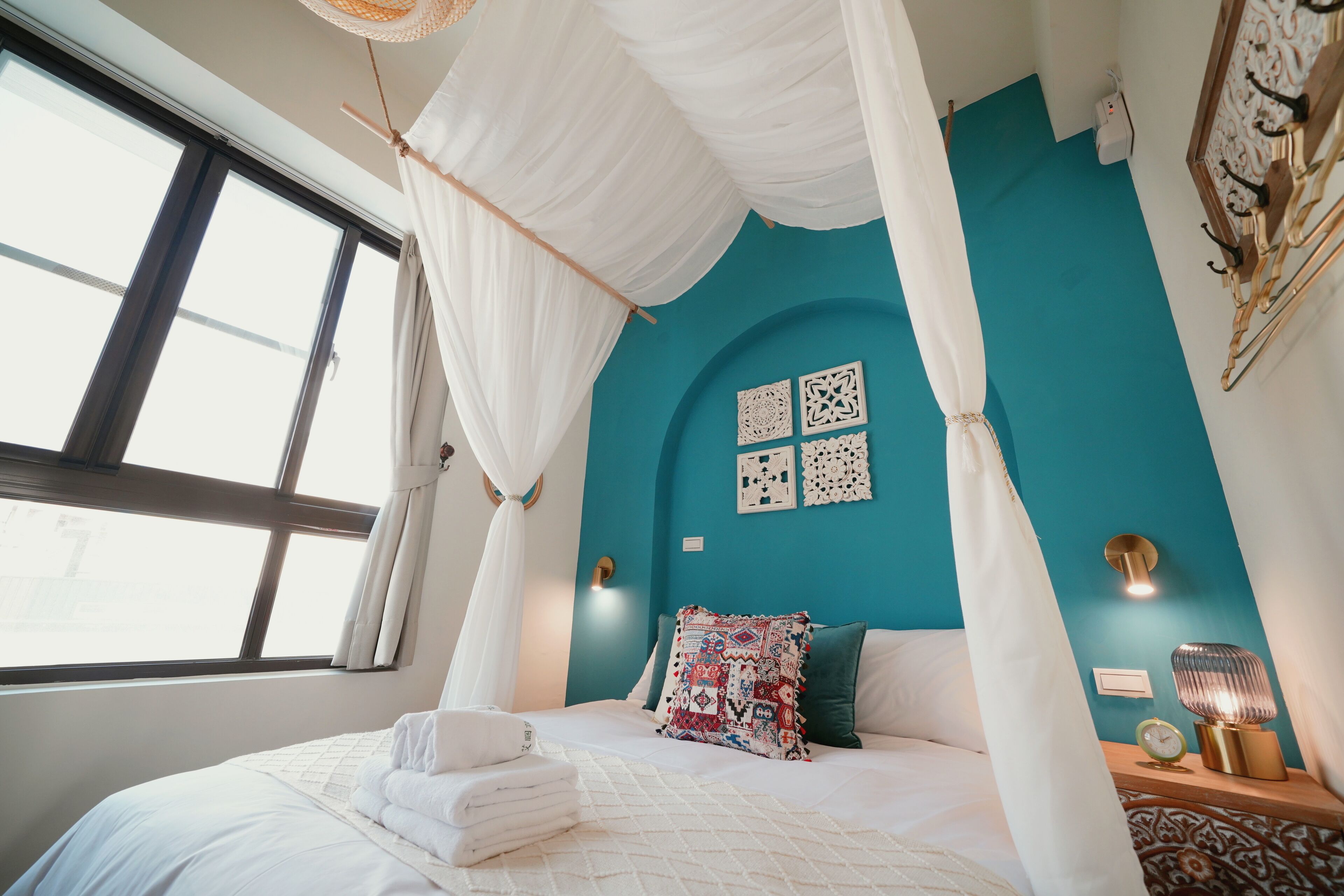 A bedroom with a bed, white curtains, a teal wall, and a nightstand with a lamp.