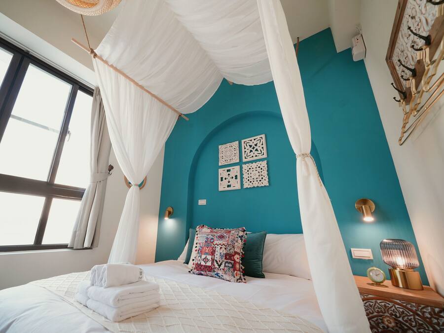 A bedroom with a bed, white curtains, a teal wall, and a nightstand with a lamp.