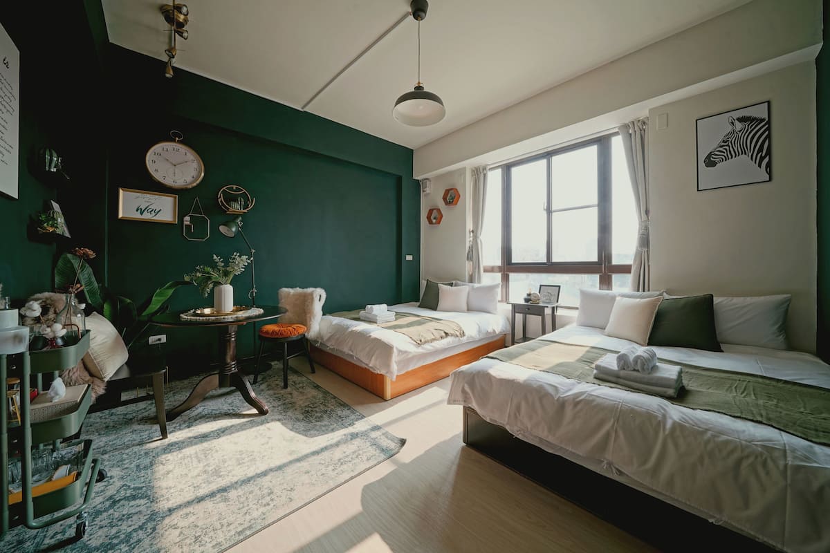 A modern bedroom with two beds, a large window, and a green accent wall.
