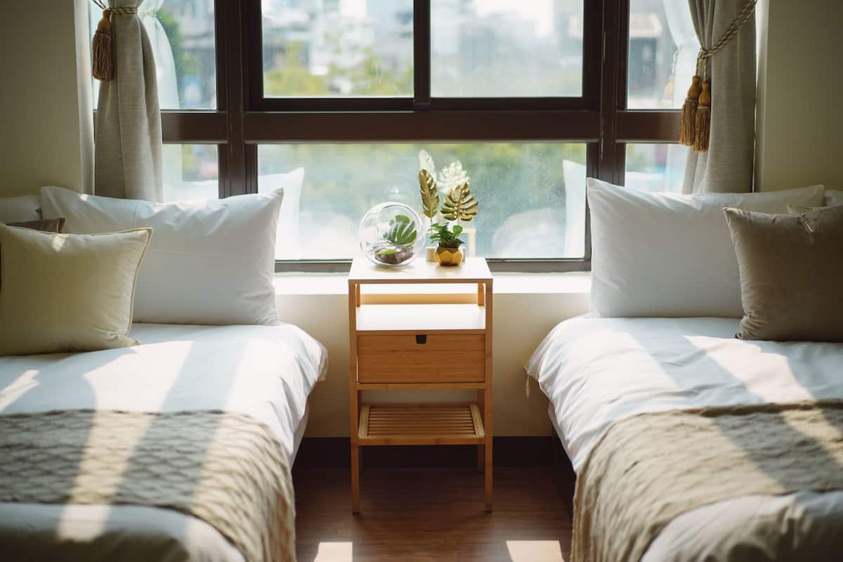 A room with two beds, a small wooden nightstand with a globe and plants, and a window with curtains.