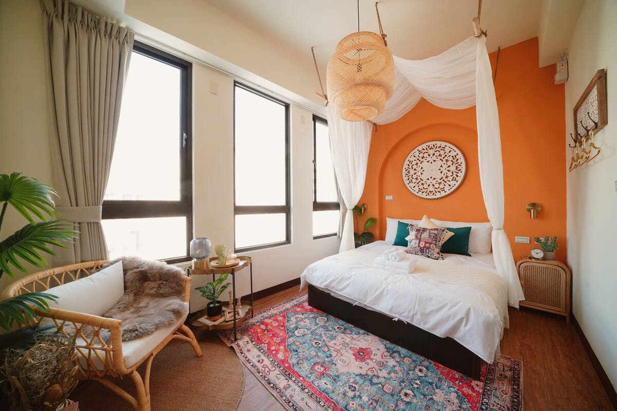 A bedroom with a large bed, a wicker chair, a small table with a plant, and a large window.