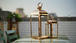 Deux lanternes de couleur dorée sont posées sur une table en verre, avec un arrière-plan extérieur flou.