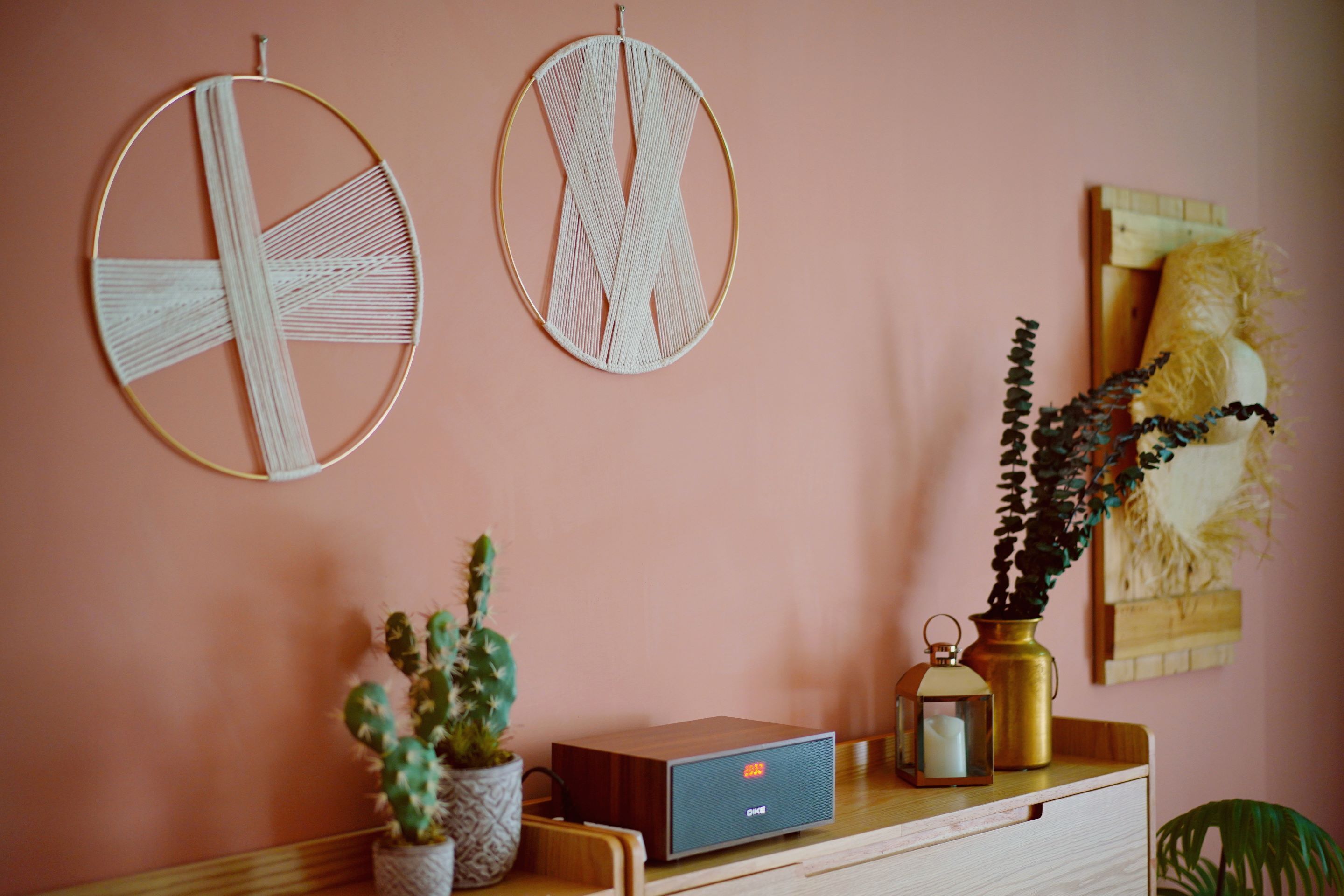 Una stanza con una parete rosa, due arazzi in macramè, un comò in legno, un cactus, una piccola pianta, un lanternino in metallo e una testiera in legno con un cuscino decorativo.