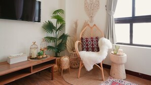 Un salon doté d’un téléviseur à écran plat, d’un banc en bois, d’un fauteuil en rotin, d’une décoration murale en macramé et d’une fenêtre avec des rideaux.