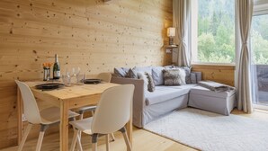 Interior de una cabaña de madera con una mesa de comedor, sillas, un sofá y una ventana que da a vistas de árboles.